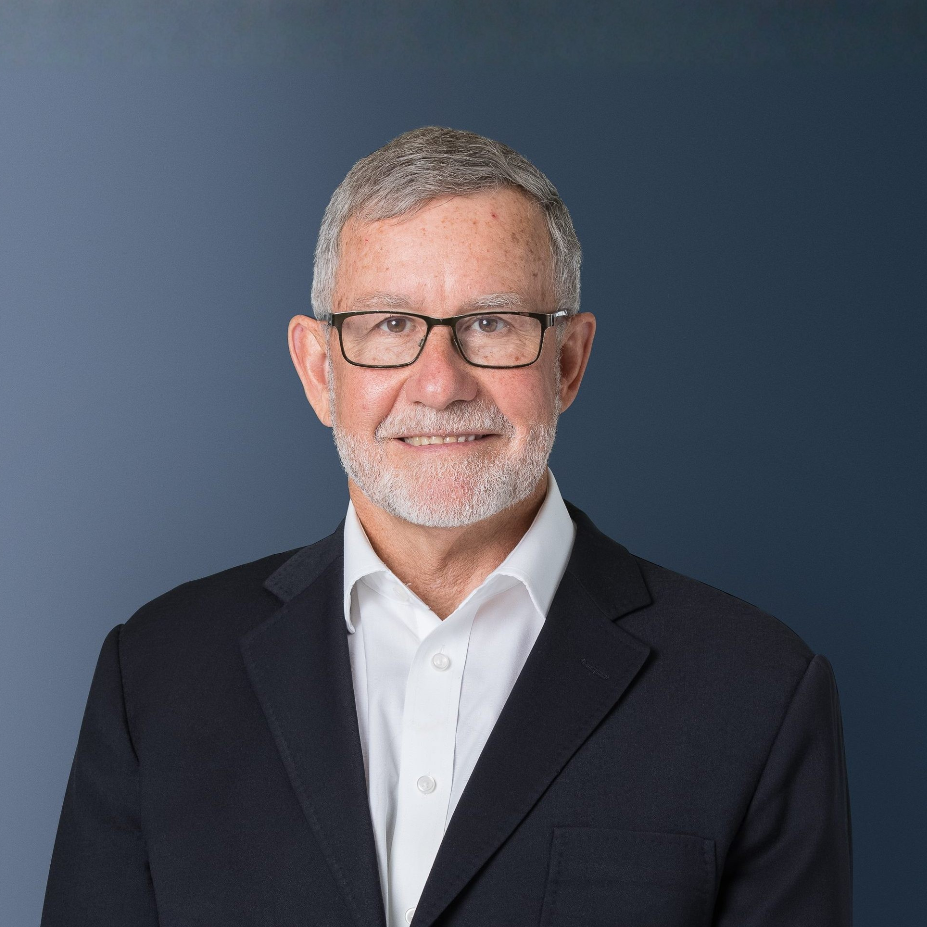 A professional portrait of an older man with gray hair, beard, and glasses, wearing a dark suit jacket and white shirt, against a plain blue background.