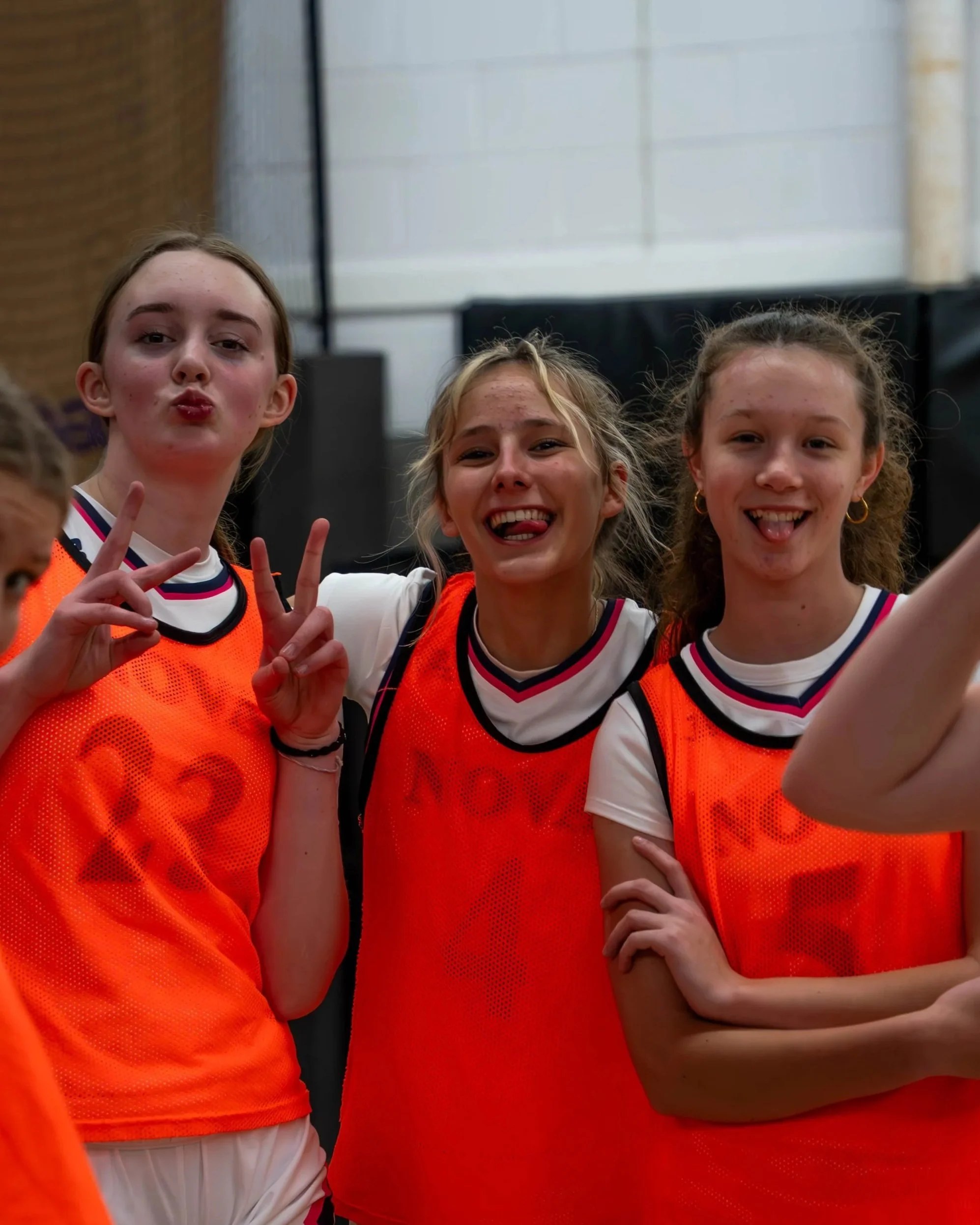 Three young girls wearing orange sports pinnies making playful and happy expressions, with one girl making a peace sign and another sticking out her tongue, in an indoor gym setting.