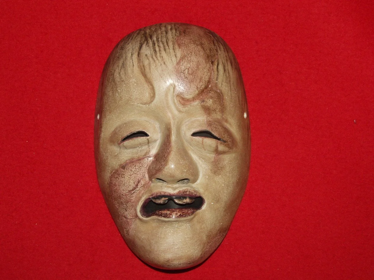 A worn, antique crying mask with distressed facial features against a red background.