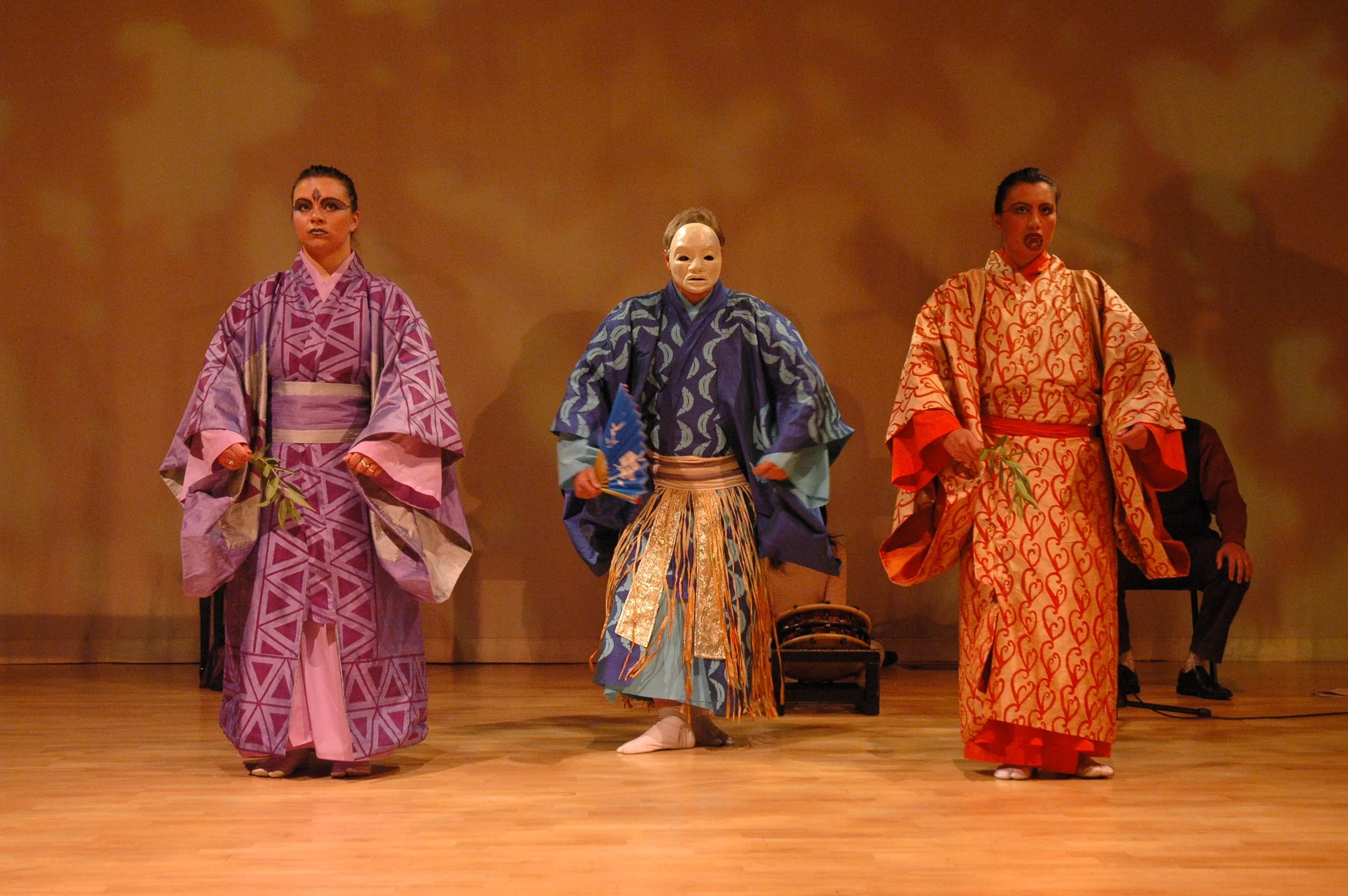 Three performers in colorful costumes on stage, holding green leaves, with a person sitting in the background.