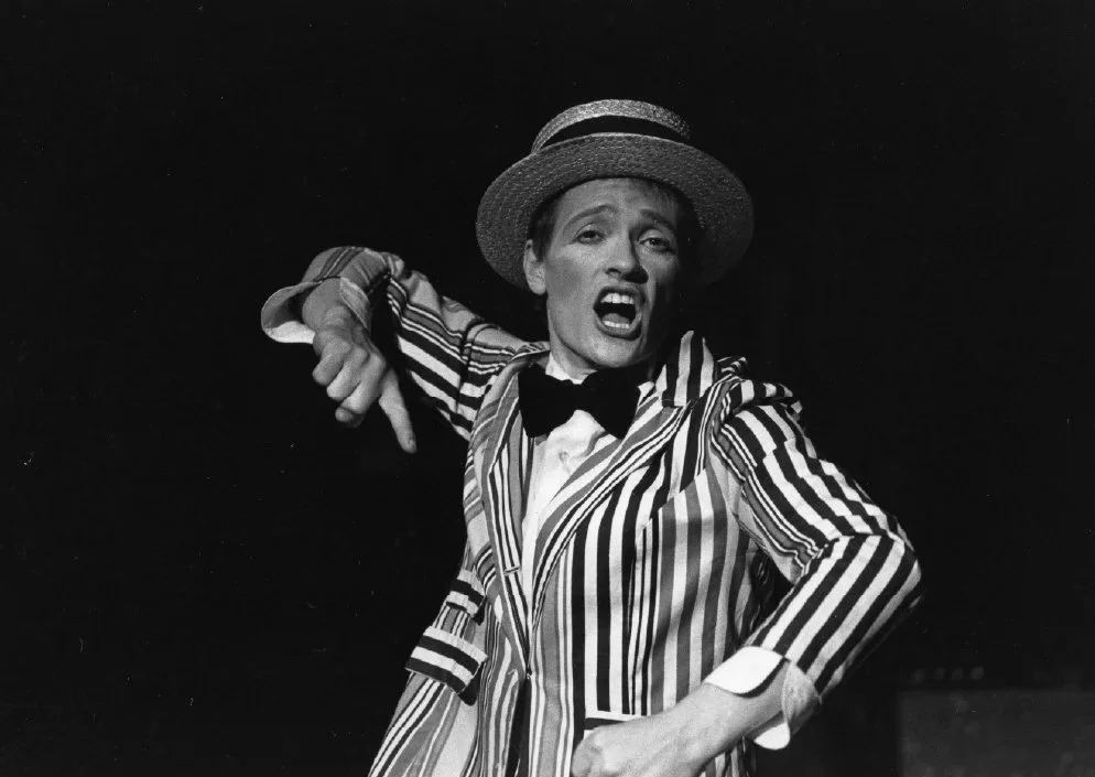 A performer singing on stage, wearing a striped jacket, a hat, and a bow tie, with expressive facial expressions.