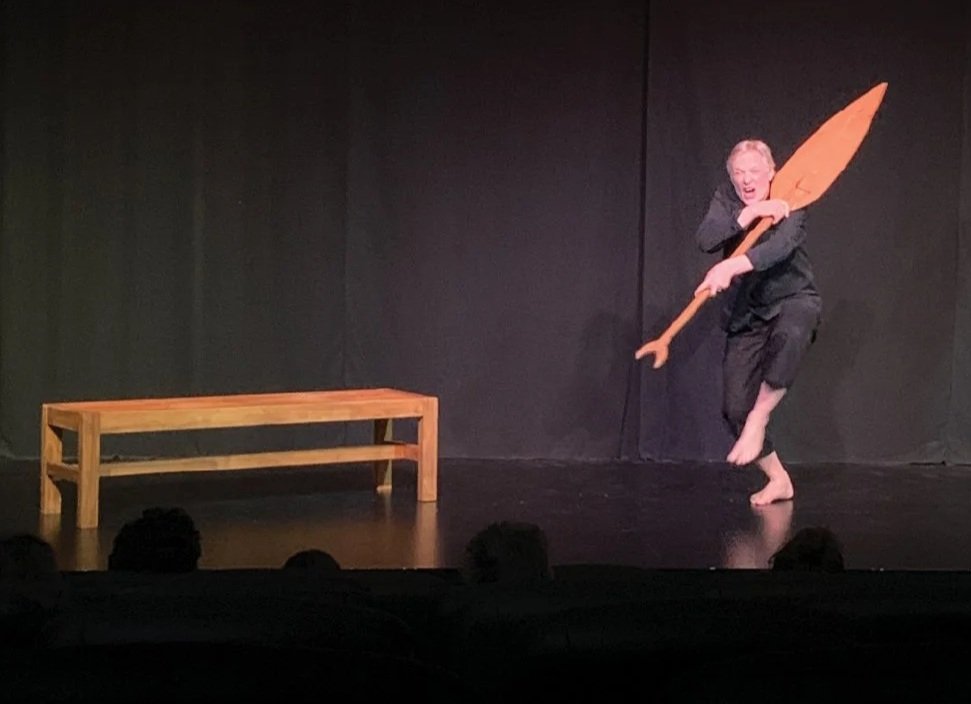 A performer on stage holding a large wooden spoon and a fork, with a wooden bench nearby, against a black curtain backdrop.