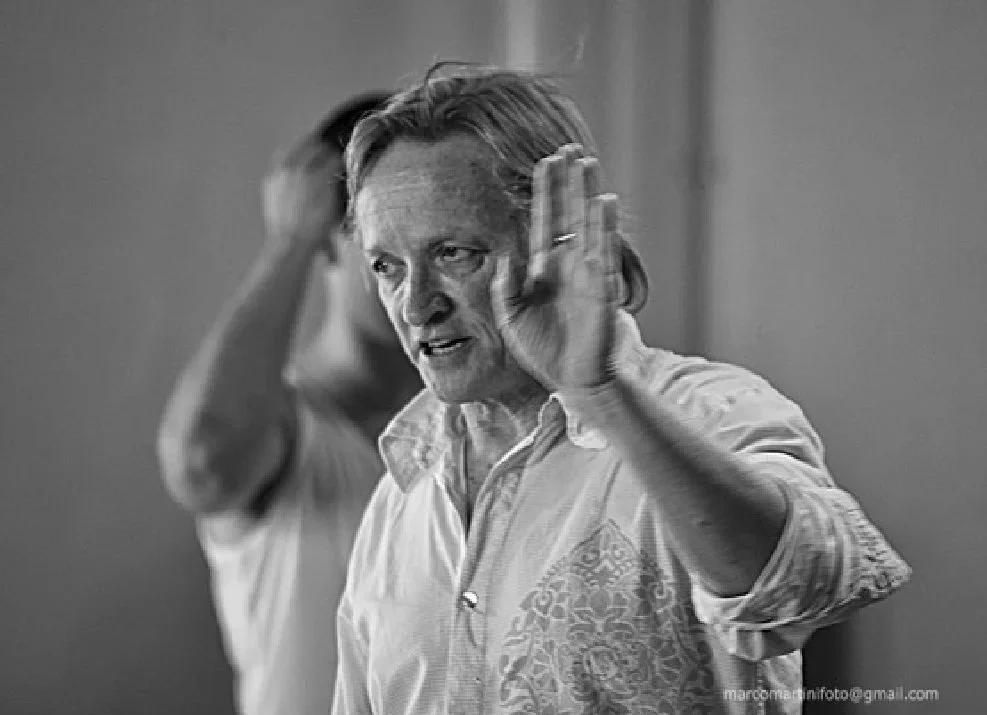 A black and white photo of a man with long hair talking, with his hands raised near his face.