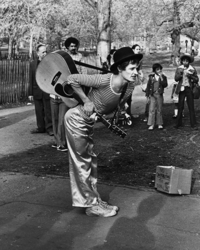 A woman dressed as a mime performing in a park, holding a guitar and bending forward, with a cardboard sign on the ground that reads 'No Imitation Too Much'. Several children and adults are watching her performance, with trees and a wooden fence in the background.