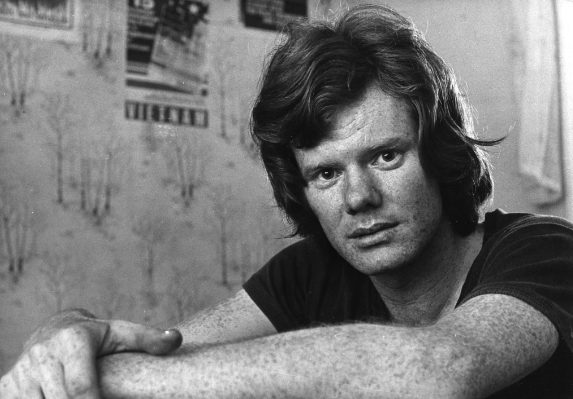Black and white photo of a young man with medium-length hair, leaning forward with his arms crossed on a surface, in a room with posters on the wall.