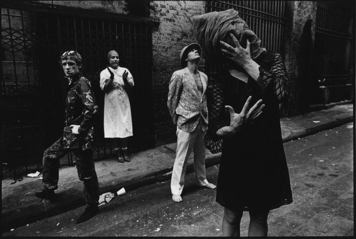 Black and white photo of four women standing on a sidewalk with a brick wall and iron fence behind them. One woman in the foreground is covering her face with her hands, wearing a headscarf and black dress. The other women are standing and watching, with one leaning against the wall.