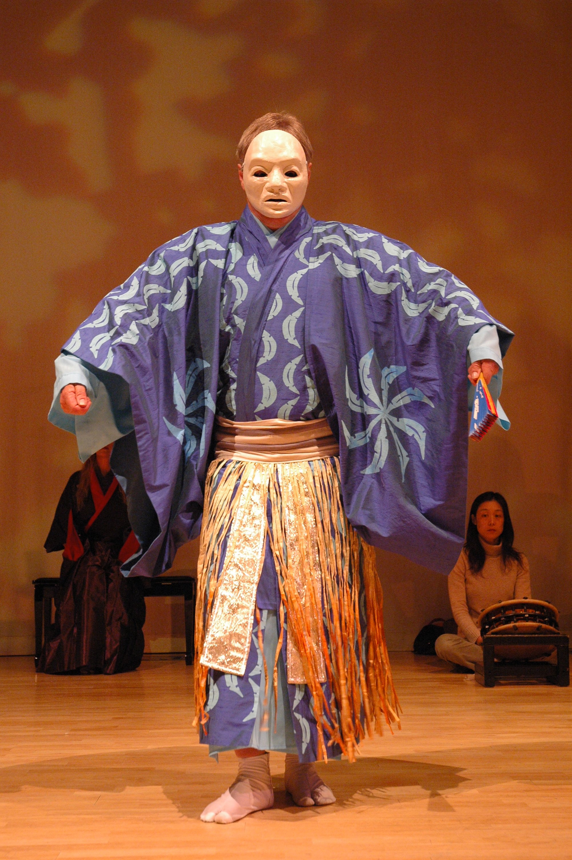 Person wearing traditional Japanese kabuki costume with a mask, standing on stage with a wooden floor and two seated individuals in the background.