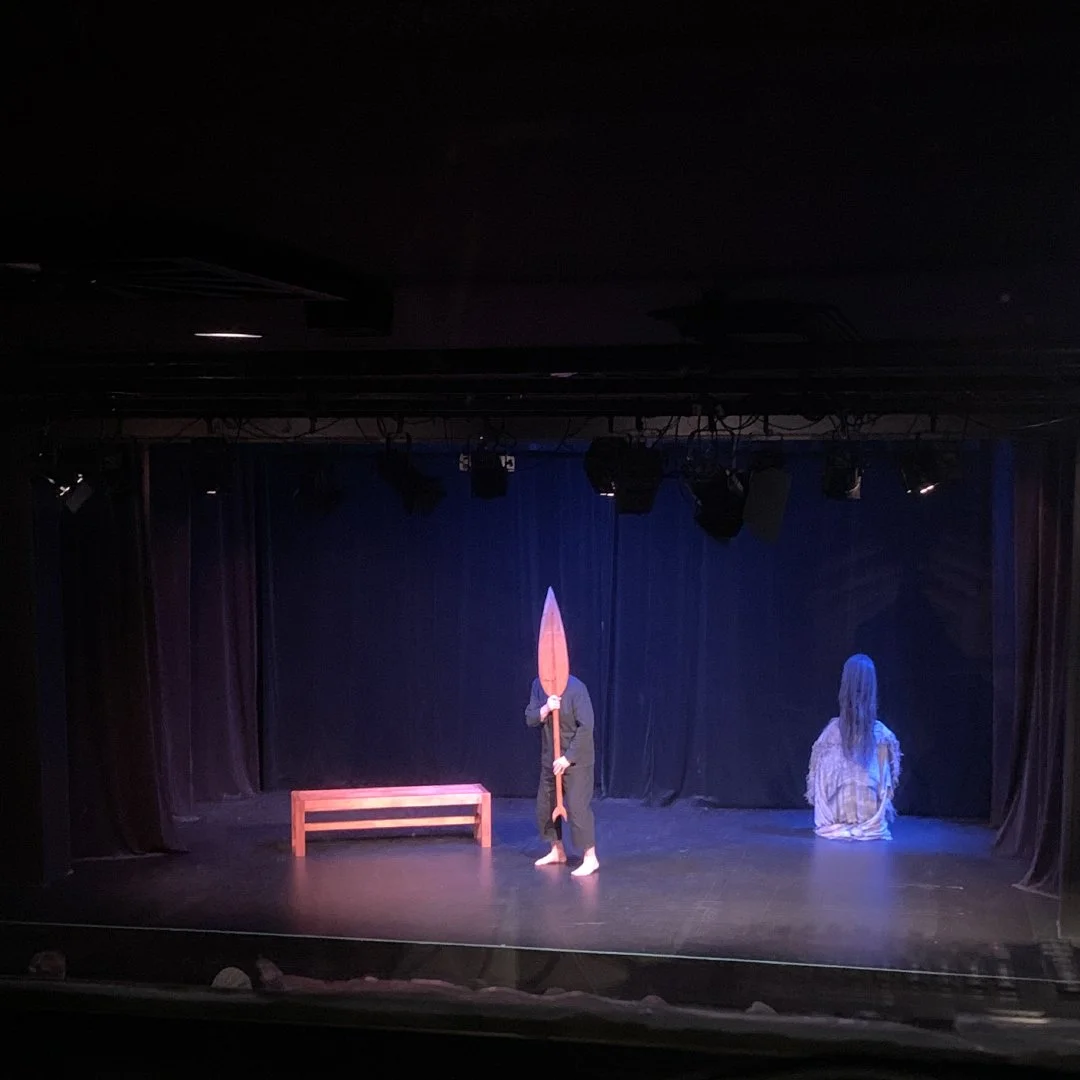 A theater stage with two performers, one holding a large wooden spear and the other sitting with long hair facing away, a bench and a black curtain backdrop, dim stage lighting.