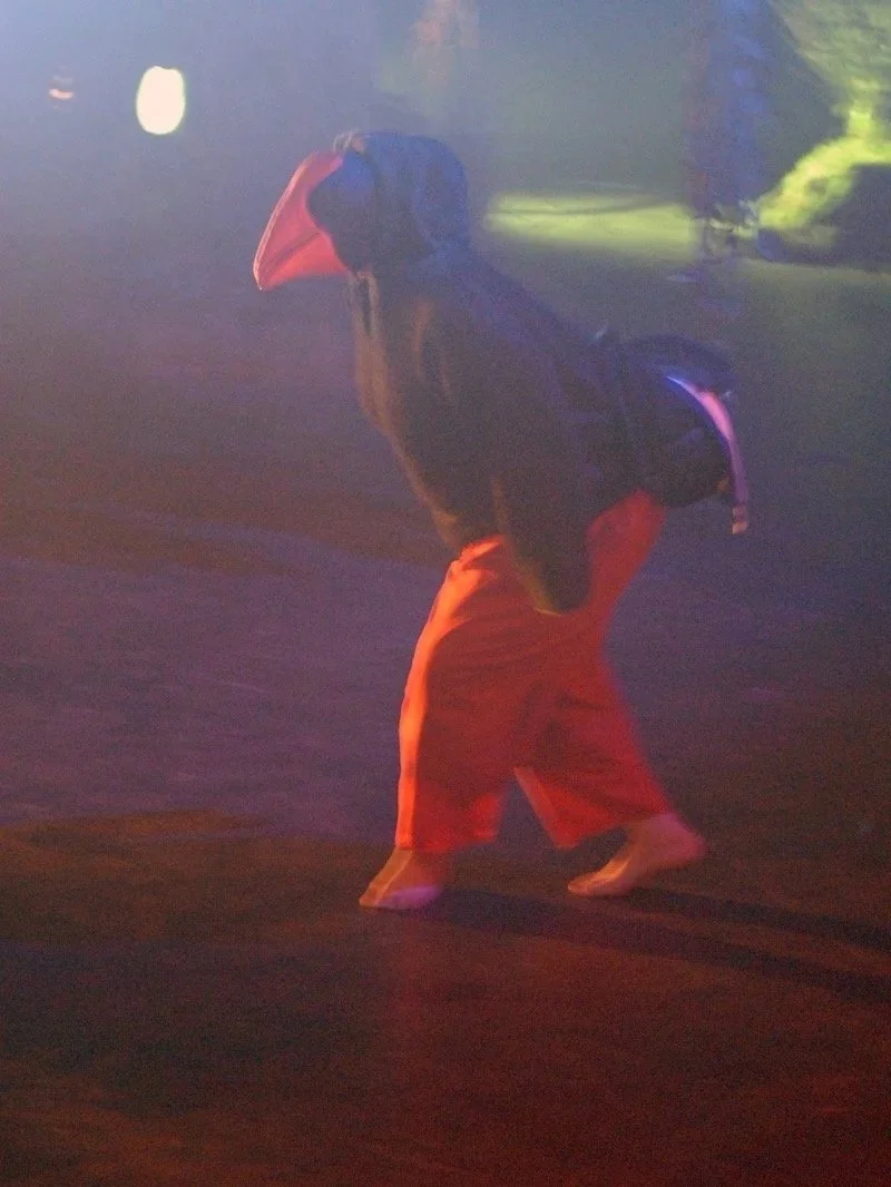 Person wearing a mohawk hairstyle, orange pants, and a dark jacket walking barefoot outdoors at night, with colorful lights illuminating the scene.