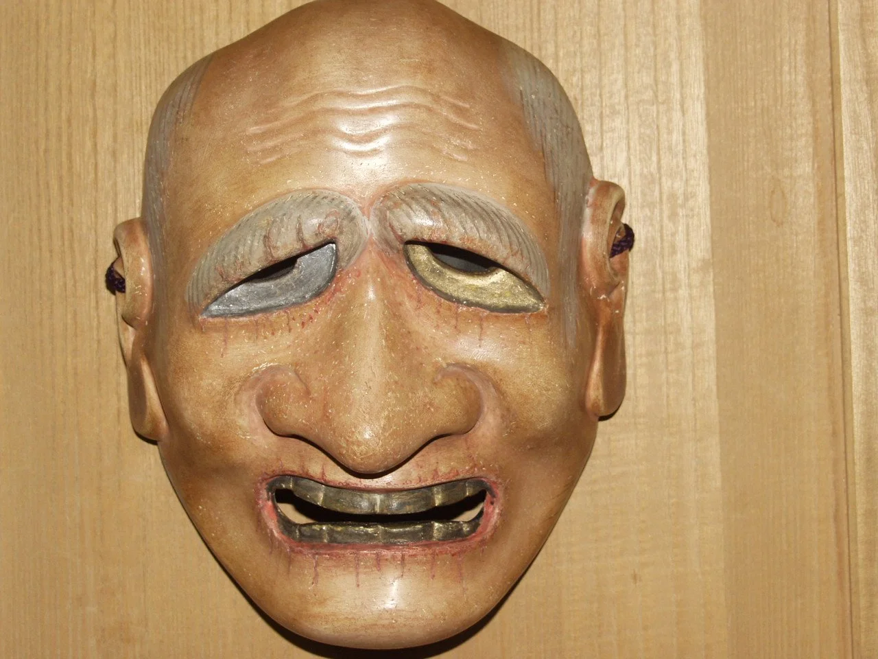 A wooden mask with a smiling face, closed eyes, prominent eyebrows, and open mouth, mounted on a wooden wall.