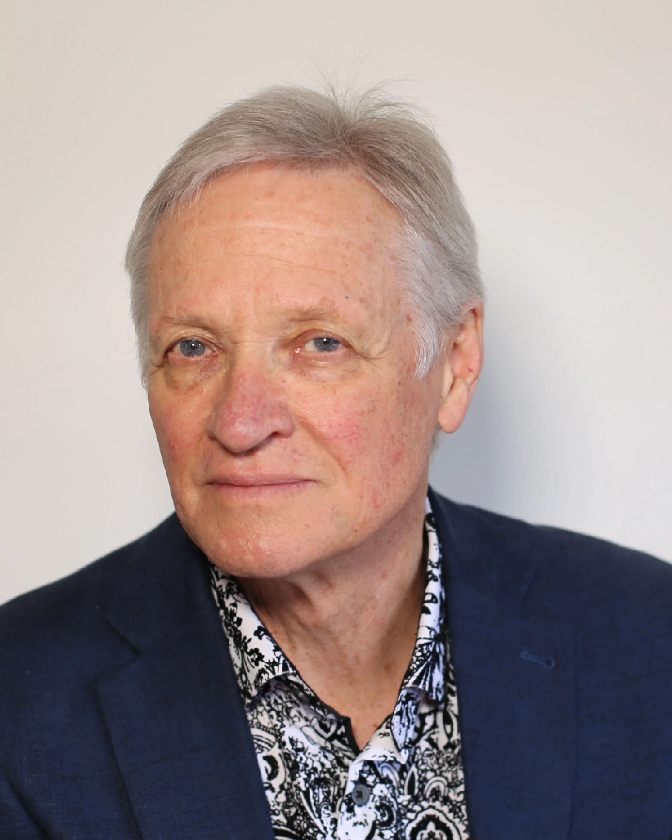 Senior man with light-colored hair and blue eyes wearing a dark blue blazer and a patterned shirt, posing against a plain light-colored background.