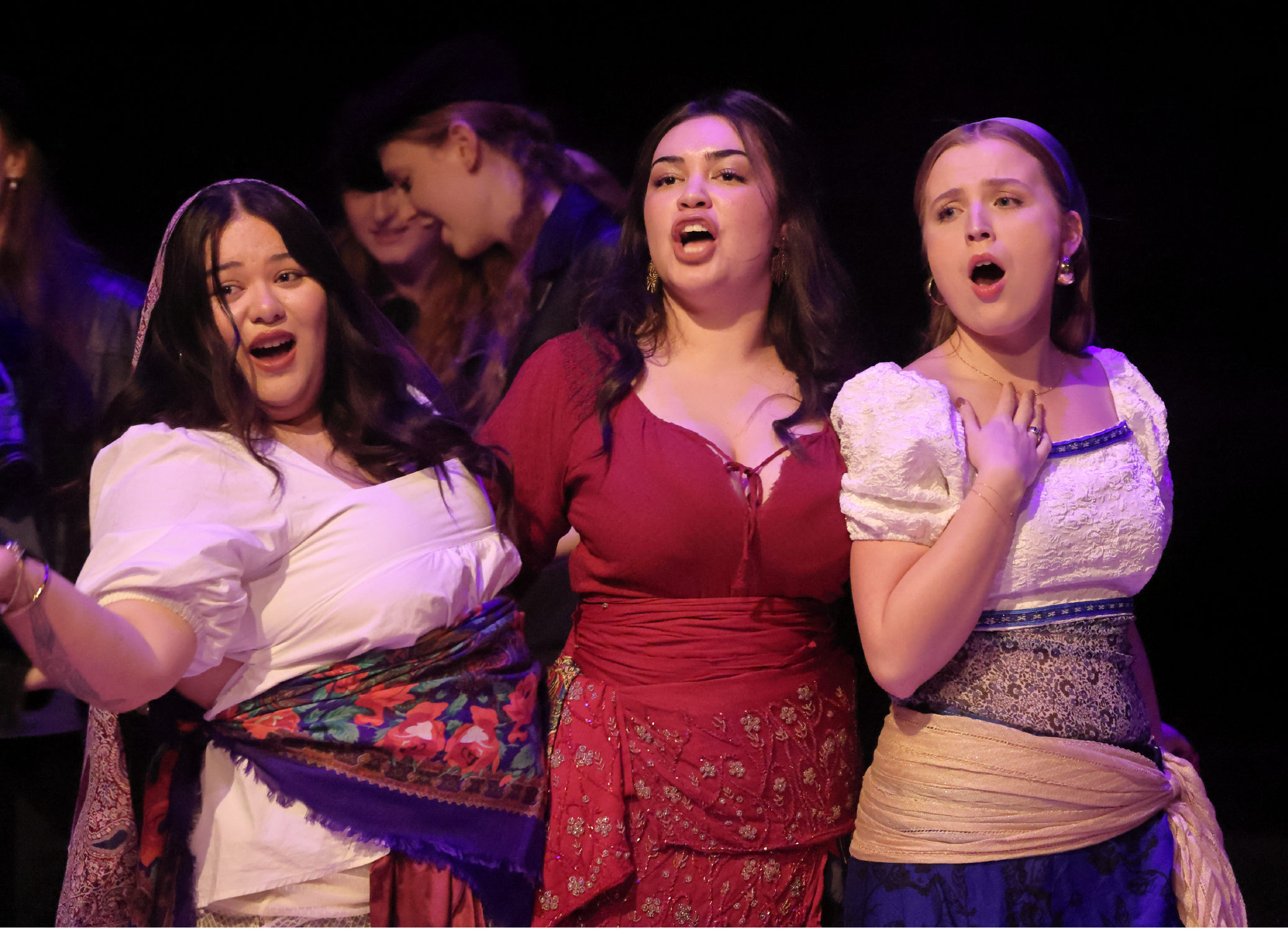 Three women singing on stage, dressed in colorful costumes, with additional women in the background.