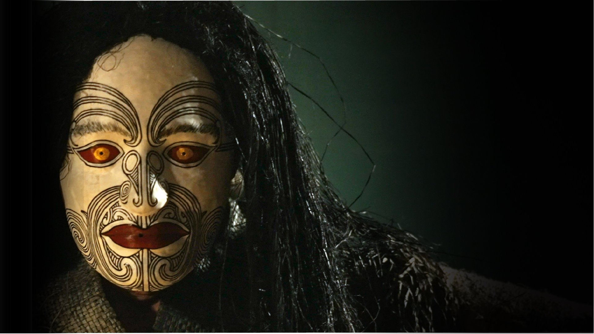 Person wearing an ornate, traditional mask with golden eyes and intricate black line patterns, with long dark hair and a dark background.