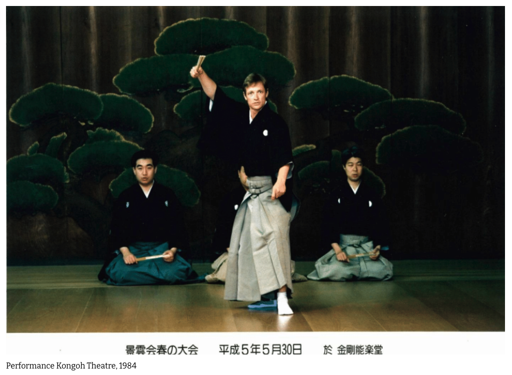 Three men dressed in traditional Japanese clothing performing a stage act at the Kongoh Theatre in 1984. The man in the center is standing and holding a knife in the air, while the other two men are sitting on the floor holding fans.