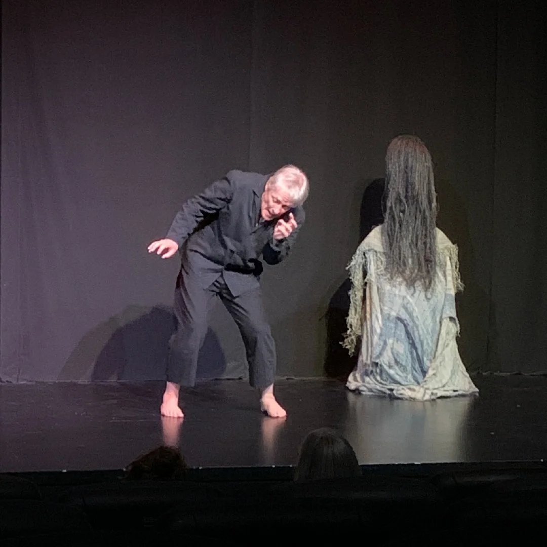A theatrical stage scene featuring an elderly man with gray hair, dressed in a black suit, barefoot, leaning forward and appearing to speak into a phone. Behind him, a woman with long, gray hair, wearing a tattered, patterned dress, is kneeling with 
