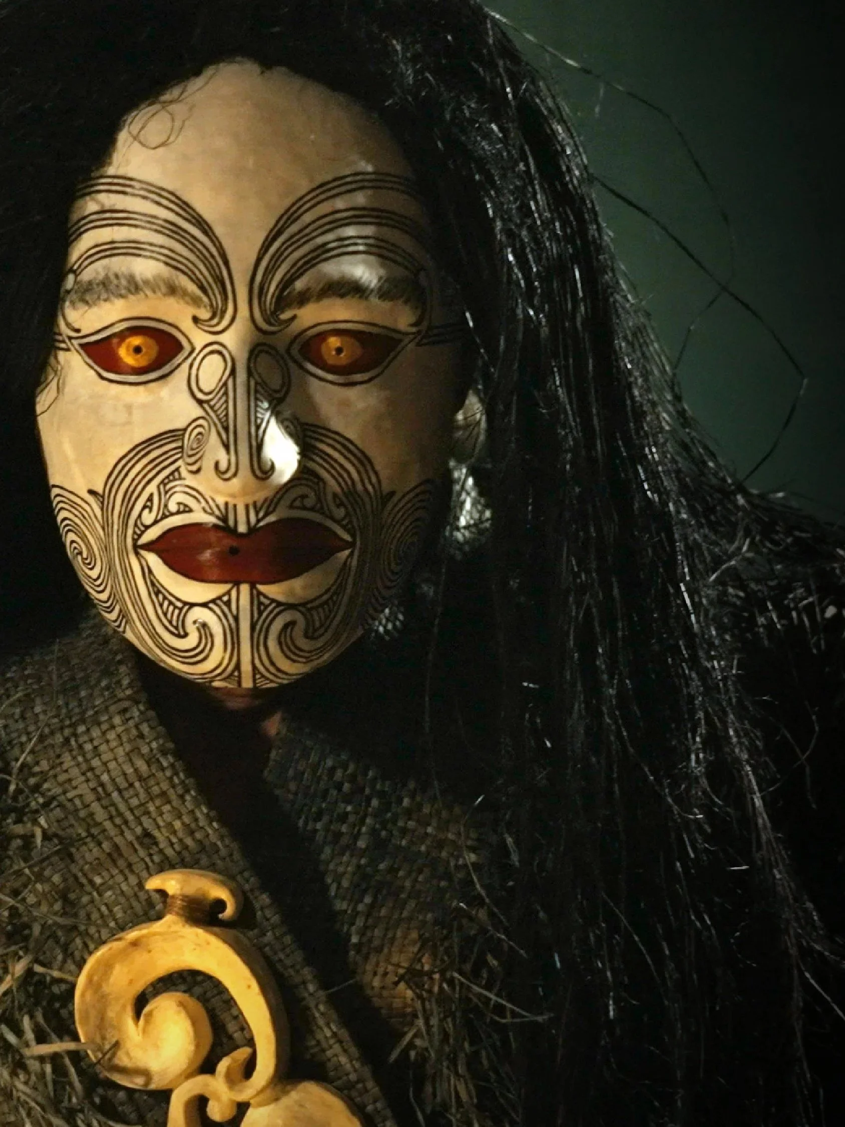 Person wearing a traditional Maori mask with intricate black line details, golden eyes, and long black hair, holding a carved bone or wood pendant, against a dark background.