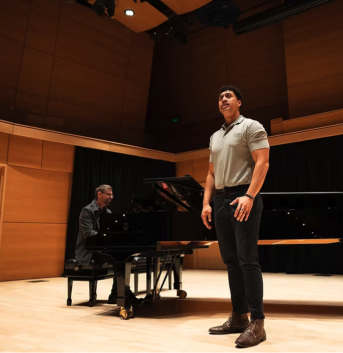 A young man singing on stage while a pianist plays a grand piano in the background.
