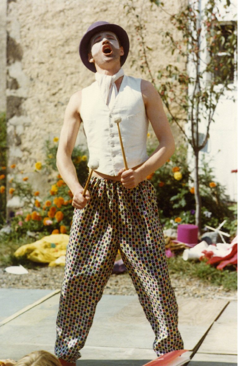 A mime performer dressed in colorful patterned pants, white vest, black hat, and white face makeup, performing outdoors in a garden with flowers and greenery.