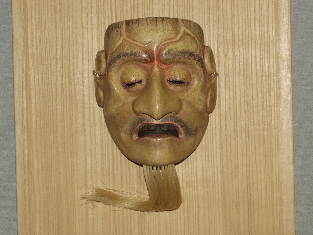 A traditional Japanese Noh mask with human facial features, including a mustache, painted with a distressed, sad expression, mounted on a wooden plaque with long, frayed hair hanging from the chin.