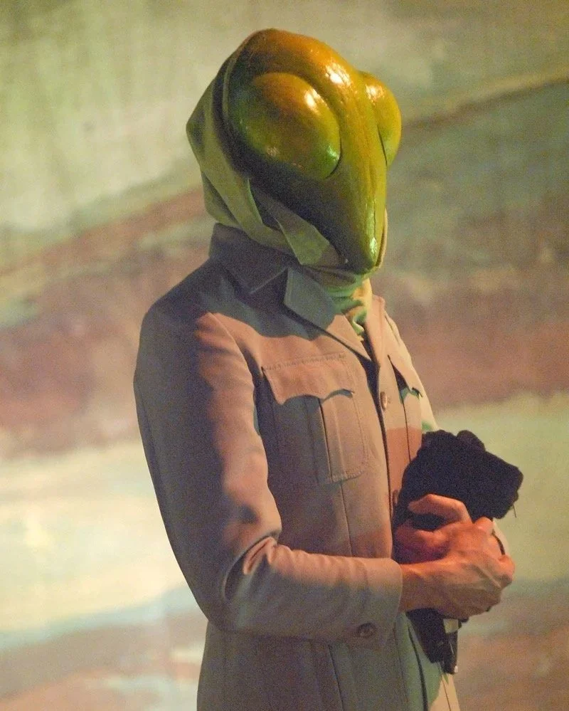 A humanoid figure with a head resembling a frog or lizard, wearing a beige jacket and holding a black item, standing in front of a yellowish background.