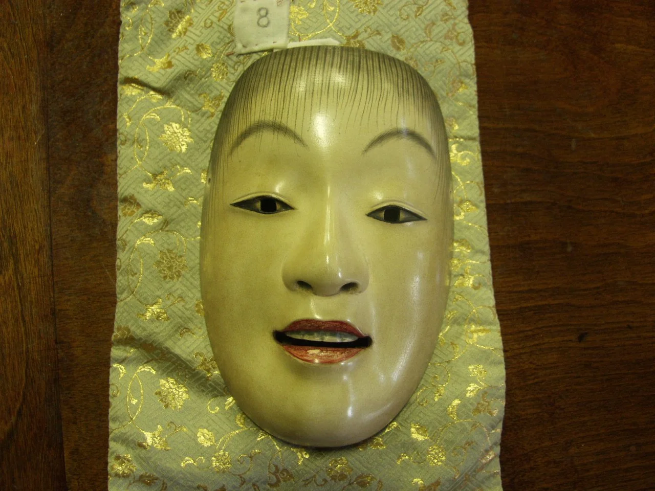 Traditional Japanese Noh mask with a serene expression, long eyebrows, small lips, and painted features, resting on a decorative cloth.