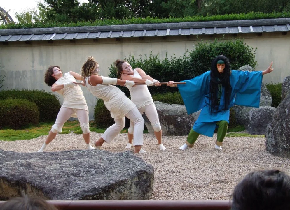 Four women are engaged in a staged performance of a tug of war against a person dressed in a traditional Japanese costume with a mask, set outdoors with rocks and greenery.