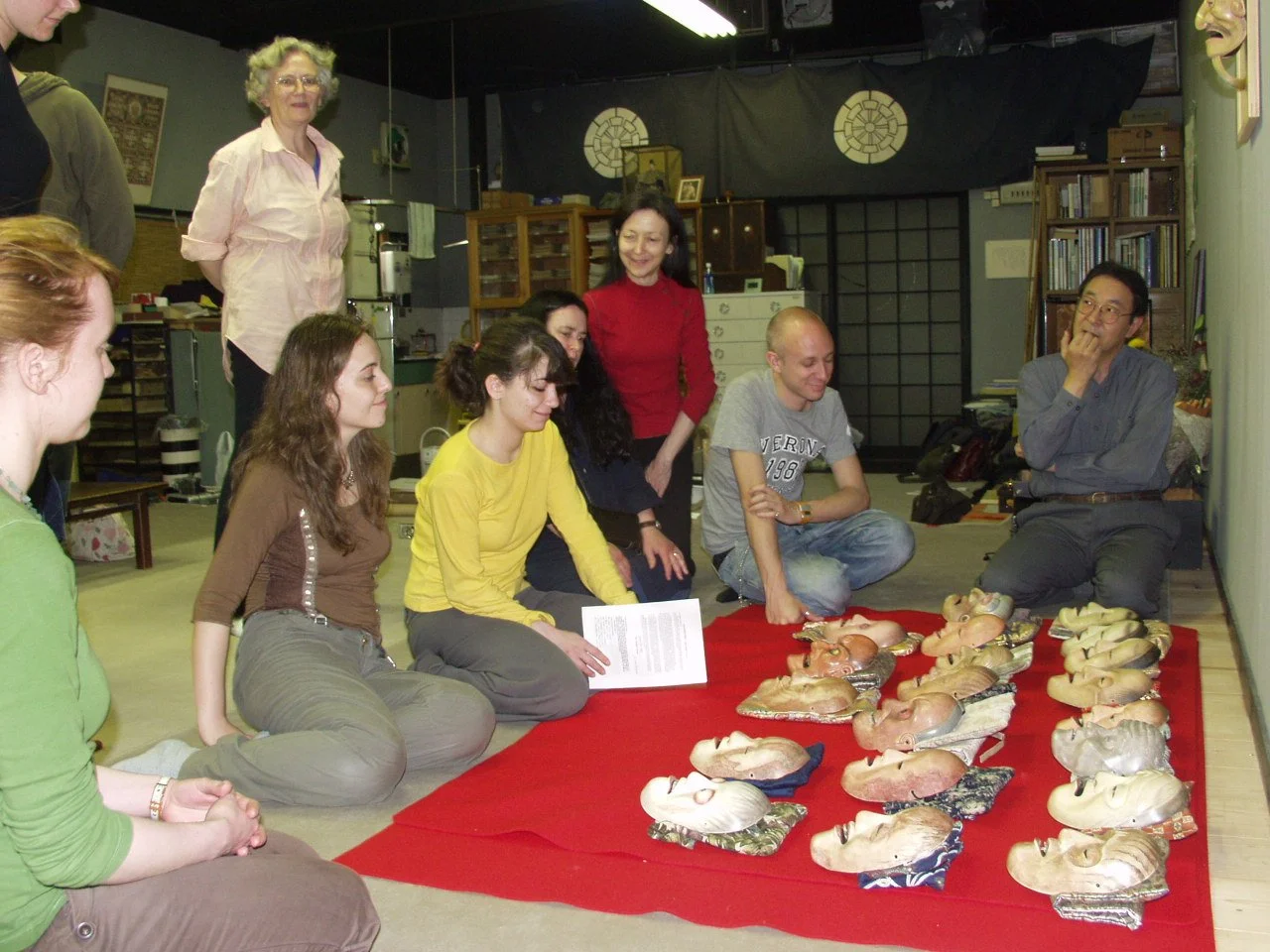 A group of people sitting and kneeling around a red cloth on the floor displaying silicone masks of various human faces. One woman stands in the background, smiling, while another woman stands near the center. The setting appears to be a room with sh