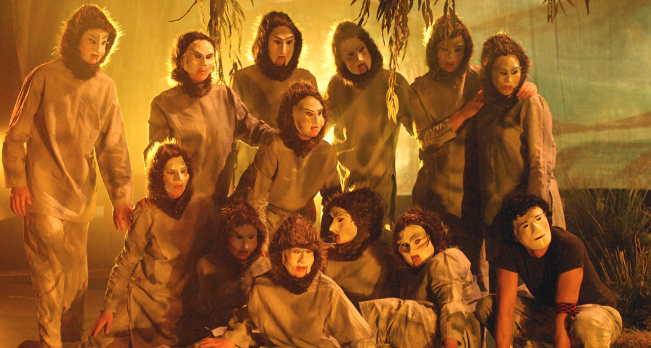 A group of performers in lion costumes and masks on a stage with dim, warm lighting and a jungle backdrop, posing together.