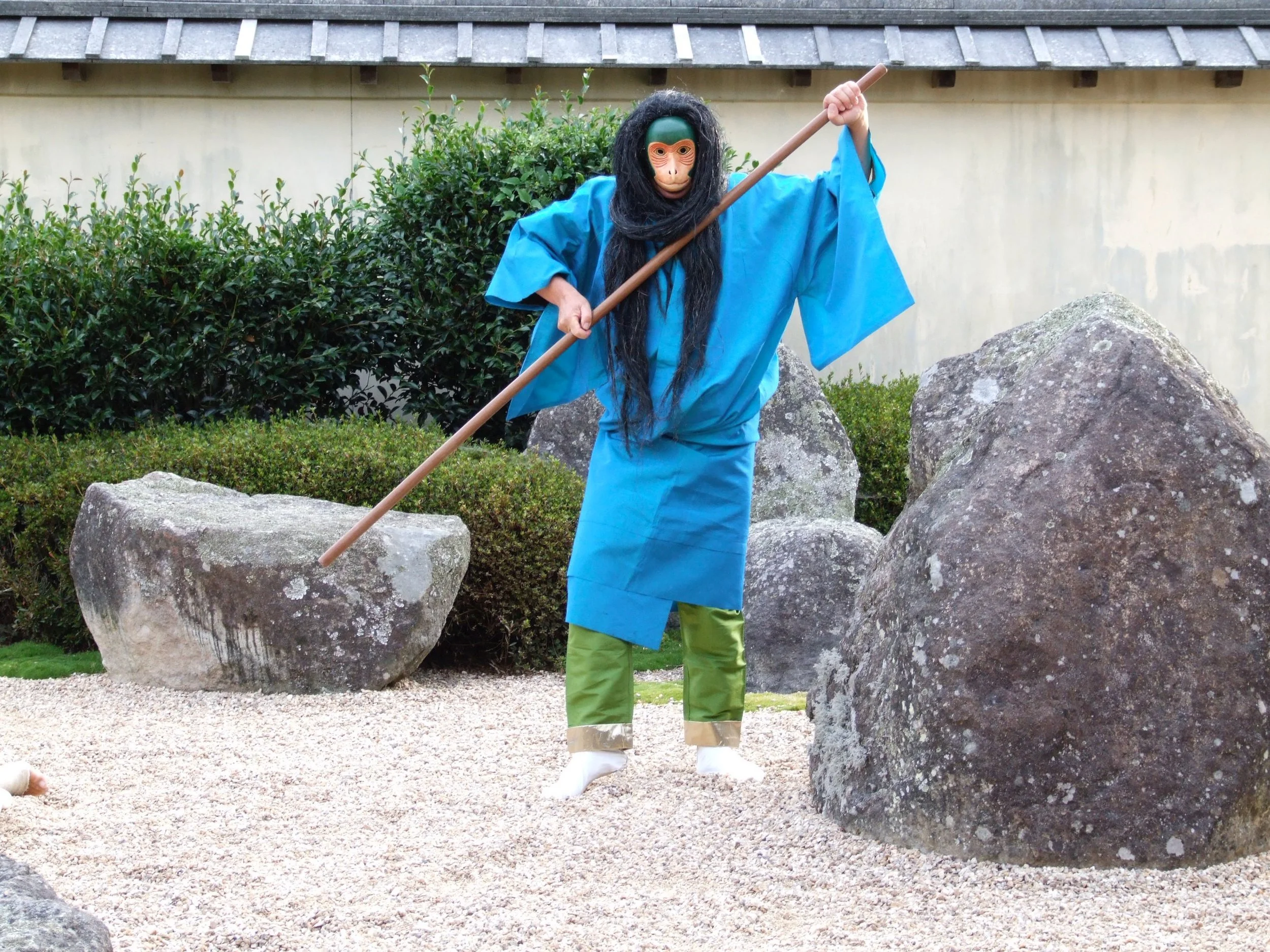 A person dressed in a monkey mask, long black hair wig, blue kimono, green pants, and white footwear holding a long stick, standing amid large rocks in a Japanese garden.