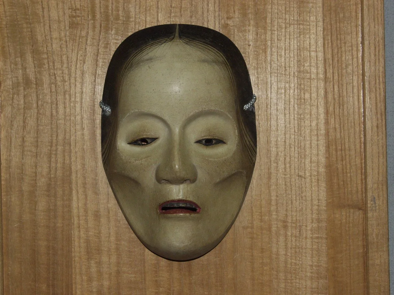 A traditional Japanese Noh mask with a somber expression, dark hair, and a beige face mounted on a wooden wall.