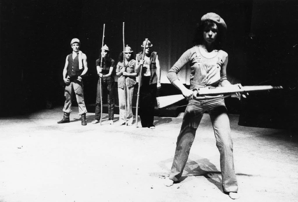 Children in a theatrical performance, with one child holding a large prop rifle in the foreground and five other children standing in a line behind, some holding sticks, on a dark stage.
