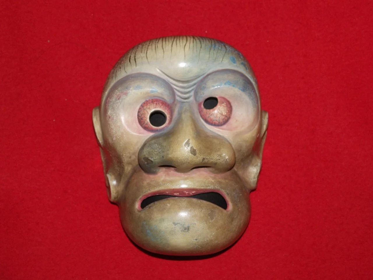 A vintage horror mask resembling a human face with exaggerated eyes, a large nose, and an open mouth, mounted on a red background.