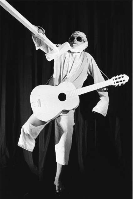 A mime performer dressed in white with sunglasses, holding a guitar and a large prop sword, standing against a dark curtain.