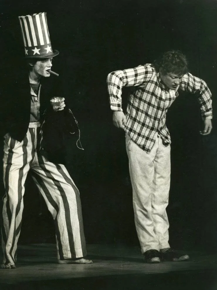 A man dressed as Uncle Sam holding a drink and cigarette, standing next to a boy with curly hair wearing a plaid shirt, on stage in a theatrical setting.