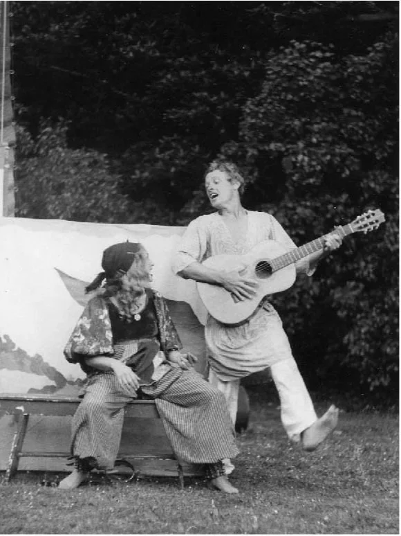 A woman playing a guitar while a clown dressed as a pirate sits nearby, outdoors at night.