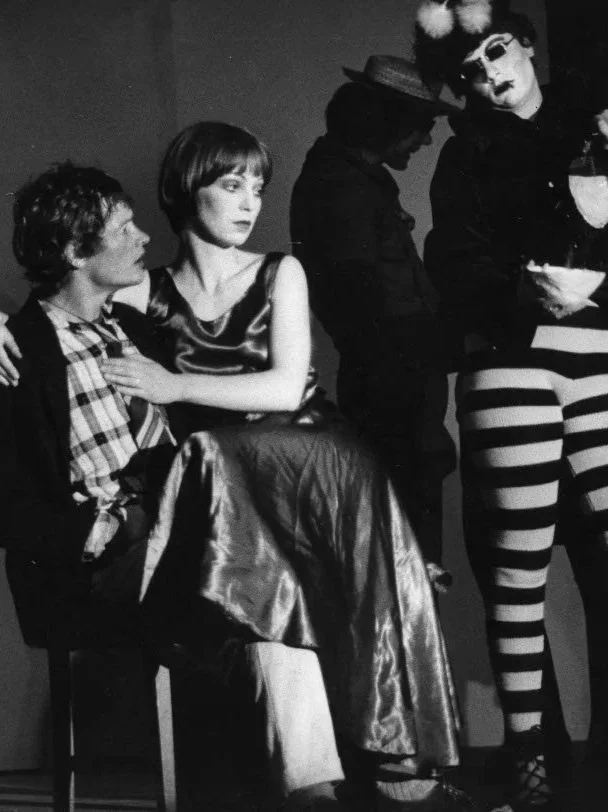 Black and white photo of four people on stage. Two seated, two standing, in theatrical costumes. The seated woman wears a shiny dress, the seated man wears a plaid shirt, and the standing person wears a dark jacket and a large hat. The other standing person is dressed as a clown with striped tights, a large wig, and dramatic makeup.
