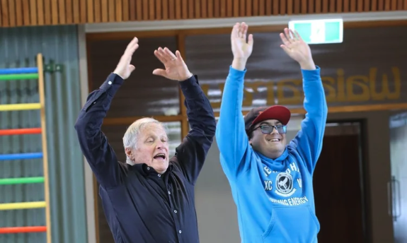An elderly woman and a young man with a cap raising their hands in celebration or victory indoors, with colorful gym equipment in the background.