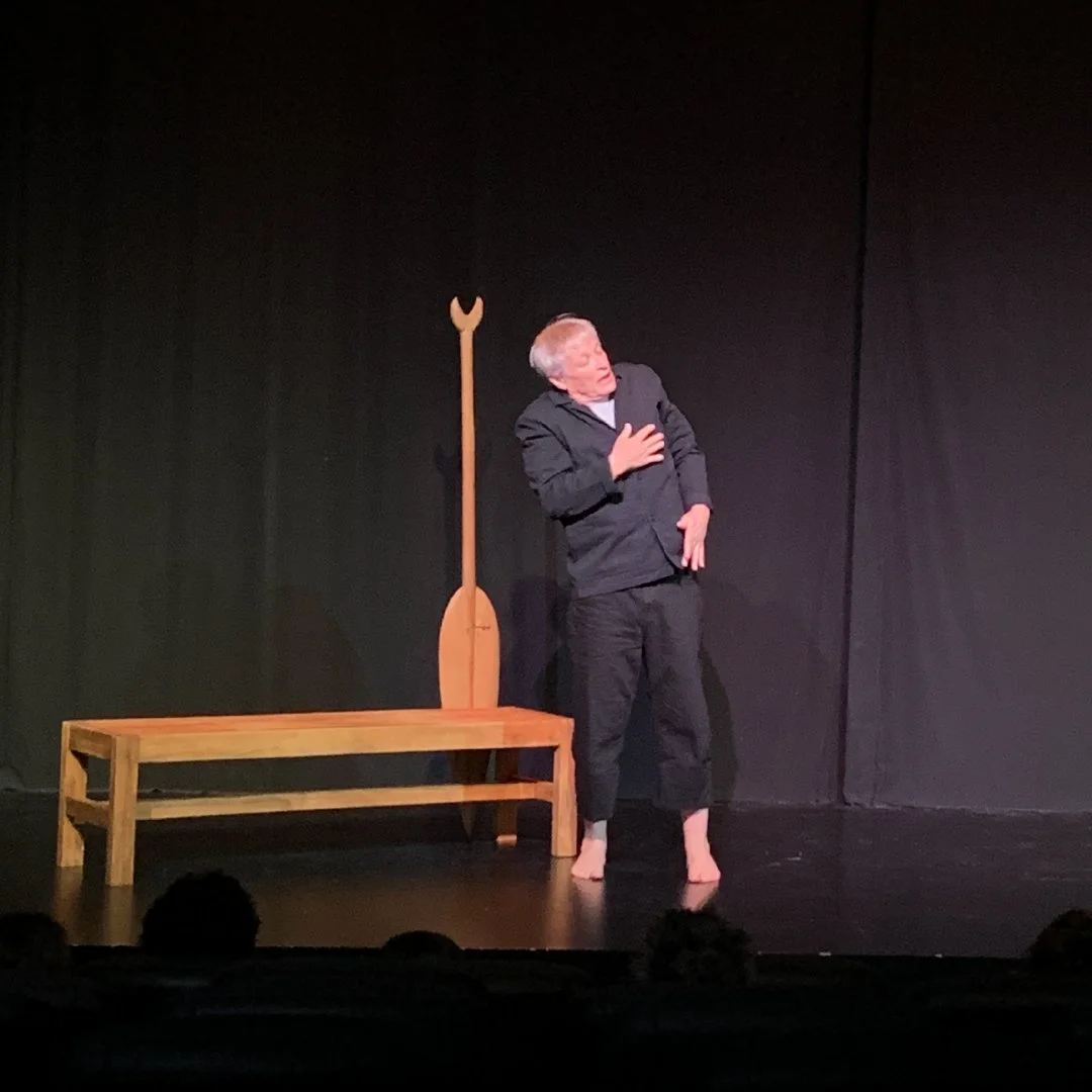 A man in a black suit is performing on stage, standing barefoot, with his hand on his chest, next to a wooden bench and a large wooden paddle with a forked top, against a black curtain backdrop.