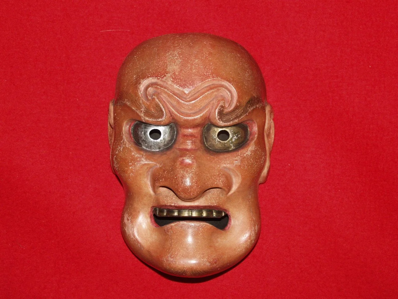 A traditional Japanese Hannya mask with a fierce, angry expression, wide eyes, and metal details, on a red background.