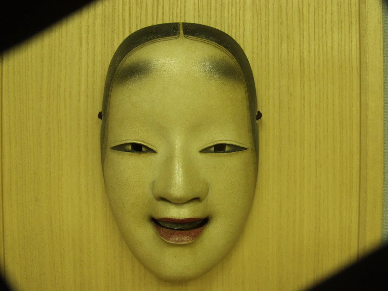 A traditional Japanese Noh mask depicting a serene, smiling woman's face with a smooth, pale complexion and black hair, mounted on a plain yellow wooden wall.