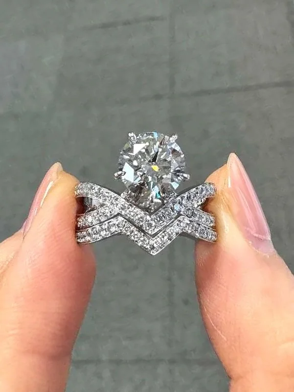 A close-up of a sparkling diamond ring with a large round diamond solitaire, held between two fingers against a plain gray background.