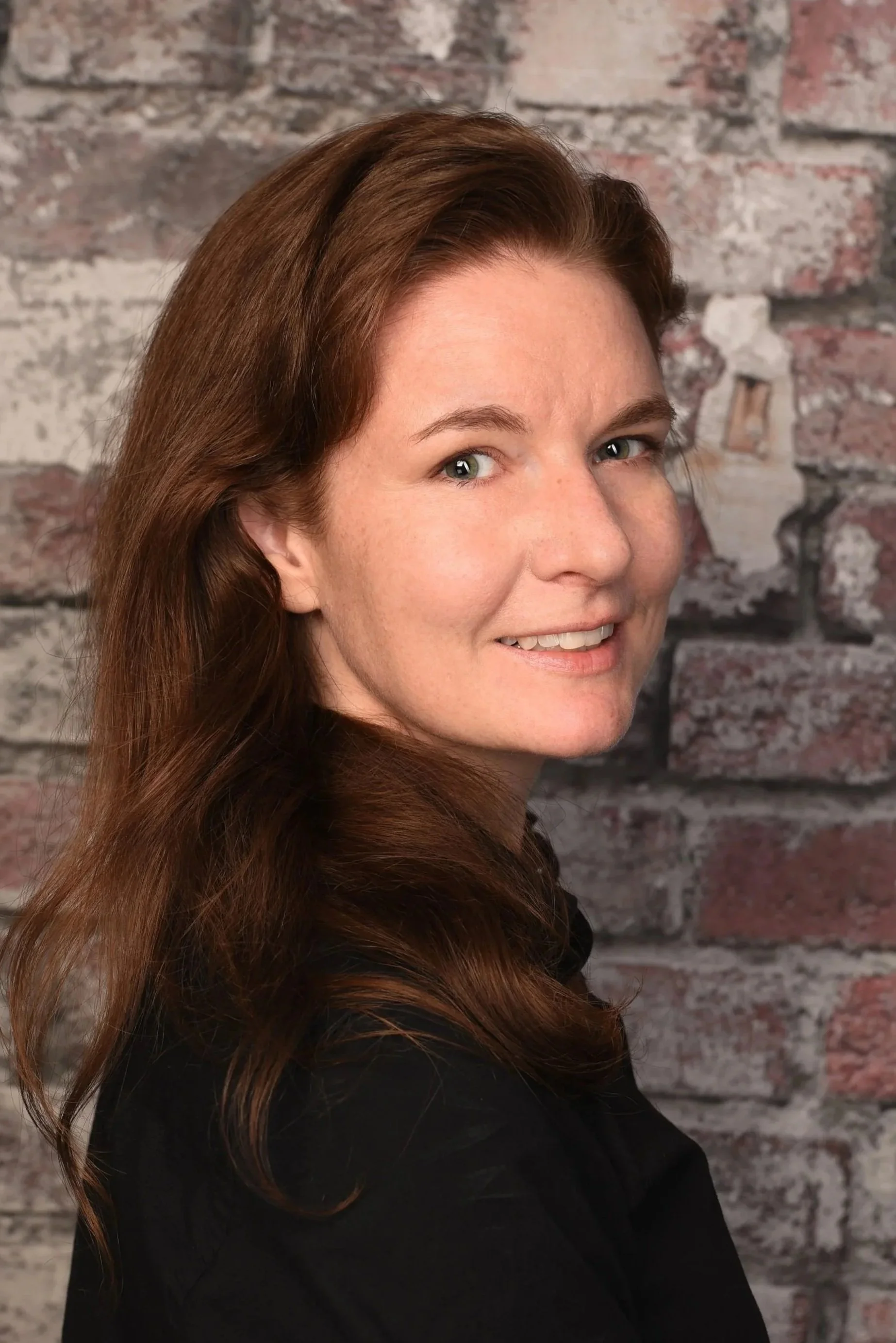 A woman with long, wavy auburn hair smiling at the camera, standing against a brick wall backdrop.