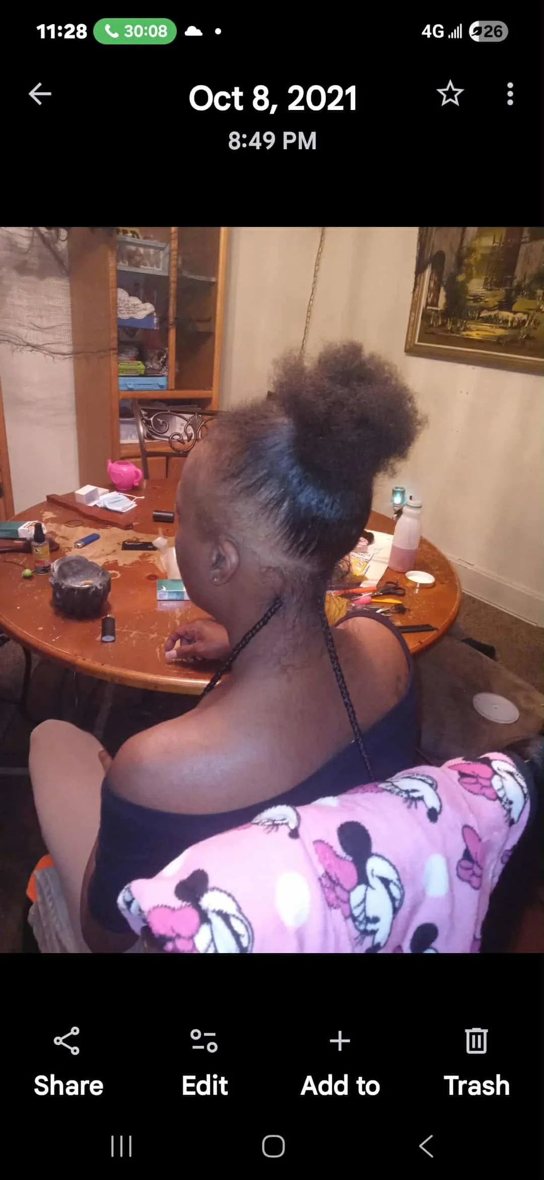 A woman with an intricate braided hairstyle, featuring a puff on top, is sitting at a dining table. The table has various hair styling tools and products on it. The woman is wearing a black off-shoulder top.