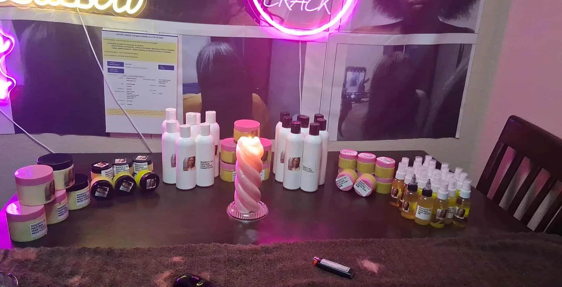 A display table with various hair care products, including bottles, jars, and small droppers, arranged in front of a mirror with pink neon lighting. There is a decorative pink candle in the center of the table, and a dark cloth covering part of the surface.