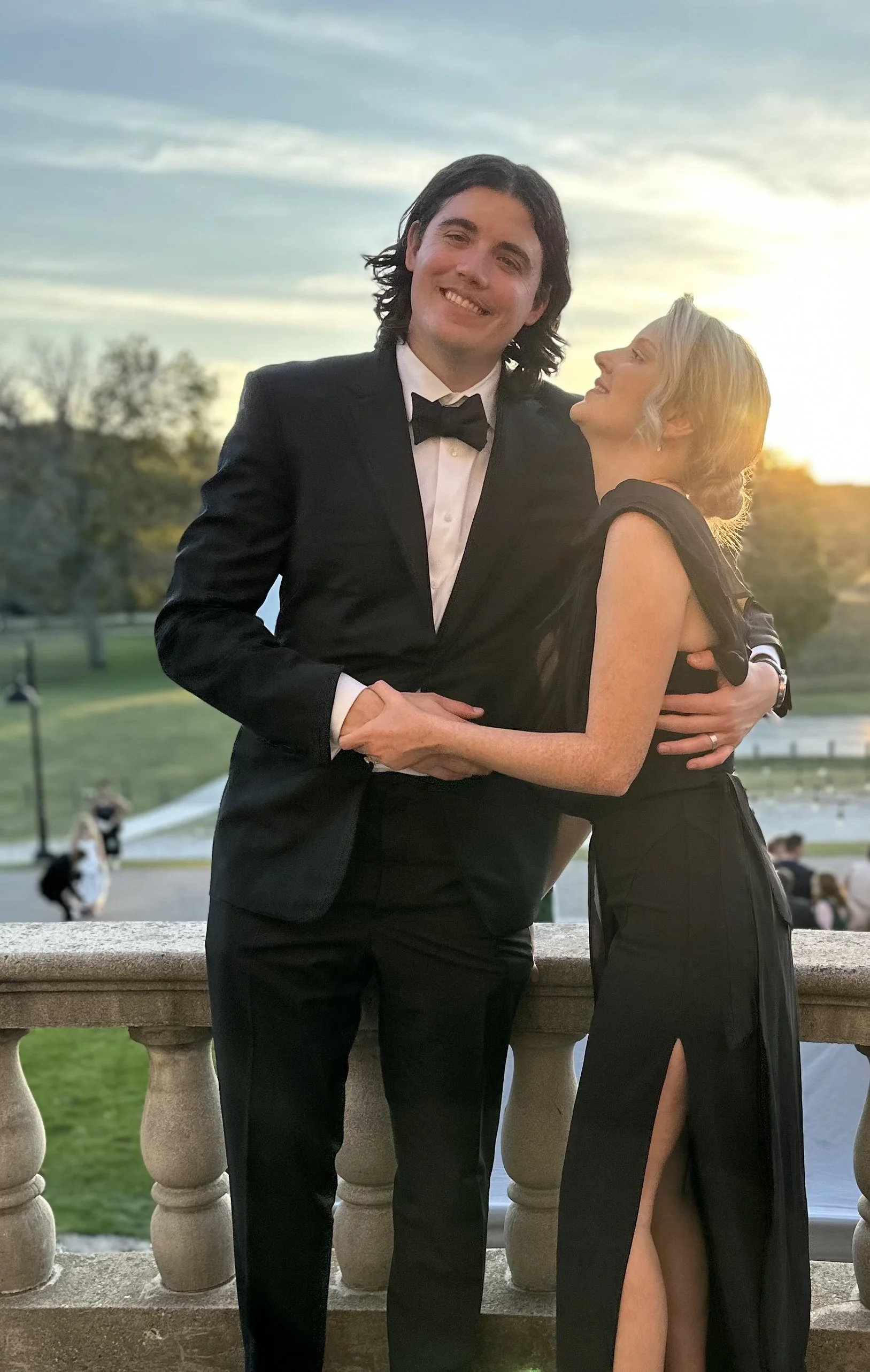 A couple dressed in formal attire, the man in a tuxedo and the woman in a black evening gown, embracing on a balcony at sunset.