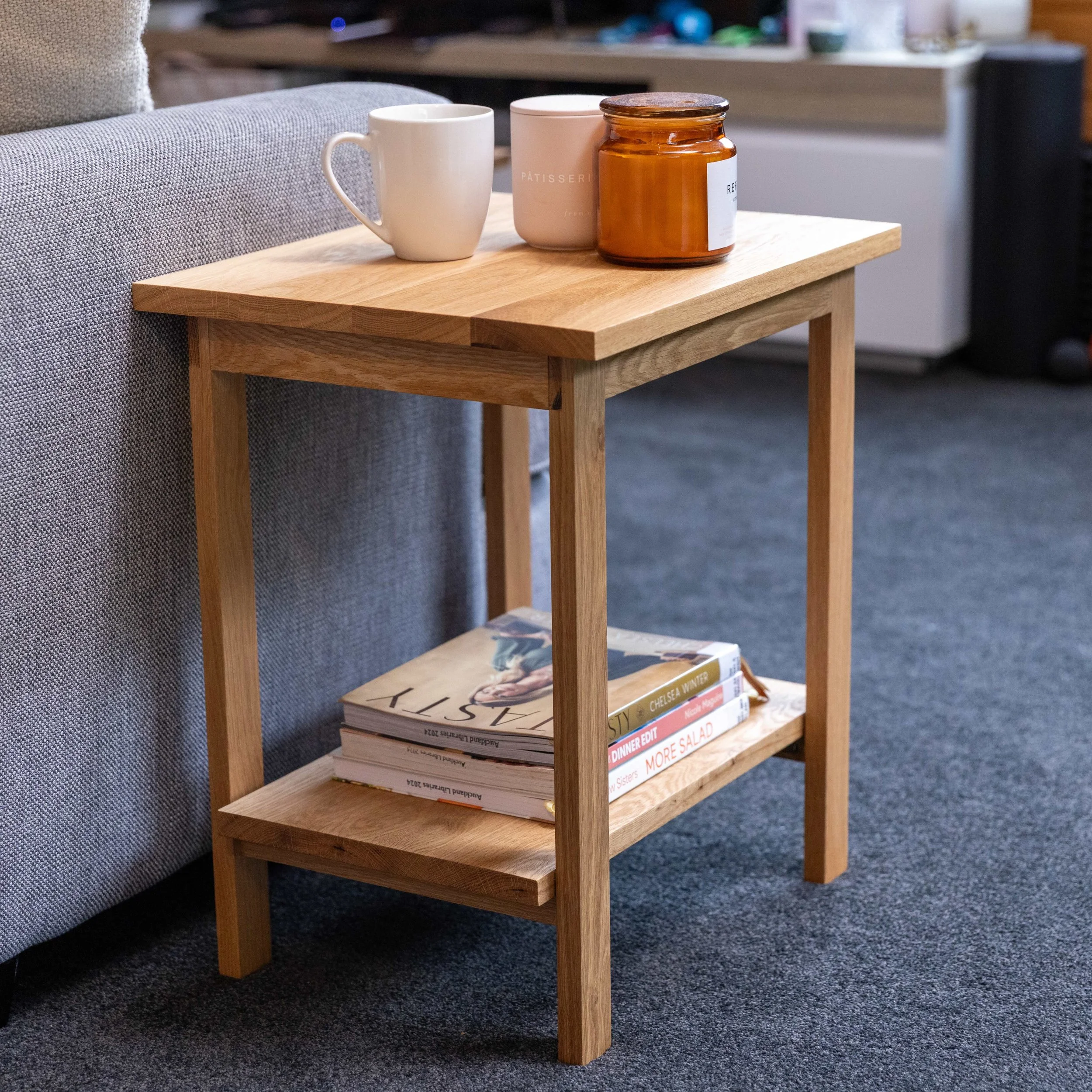 A small wooden side table next to a gray sofa, with two white mugs and a brown glass jar on top, and magazines on a lower shelf.