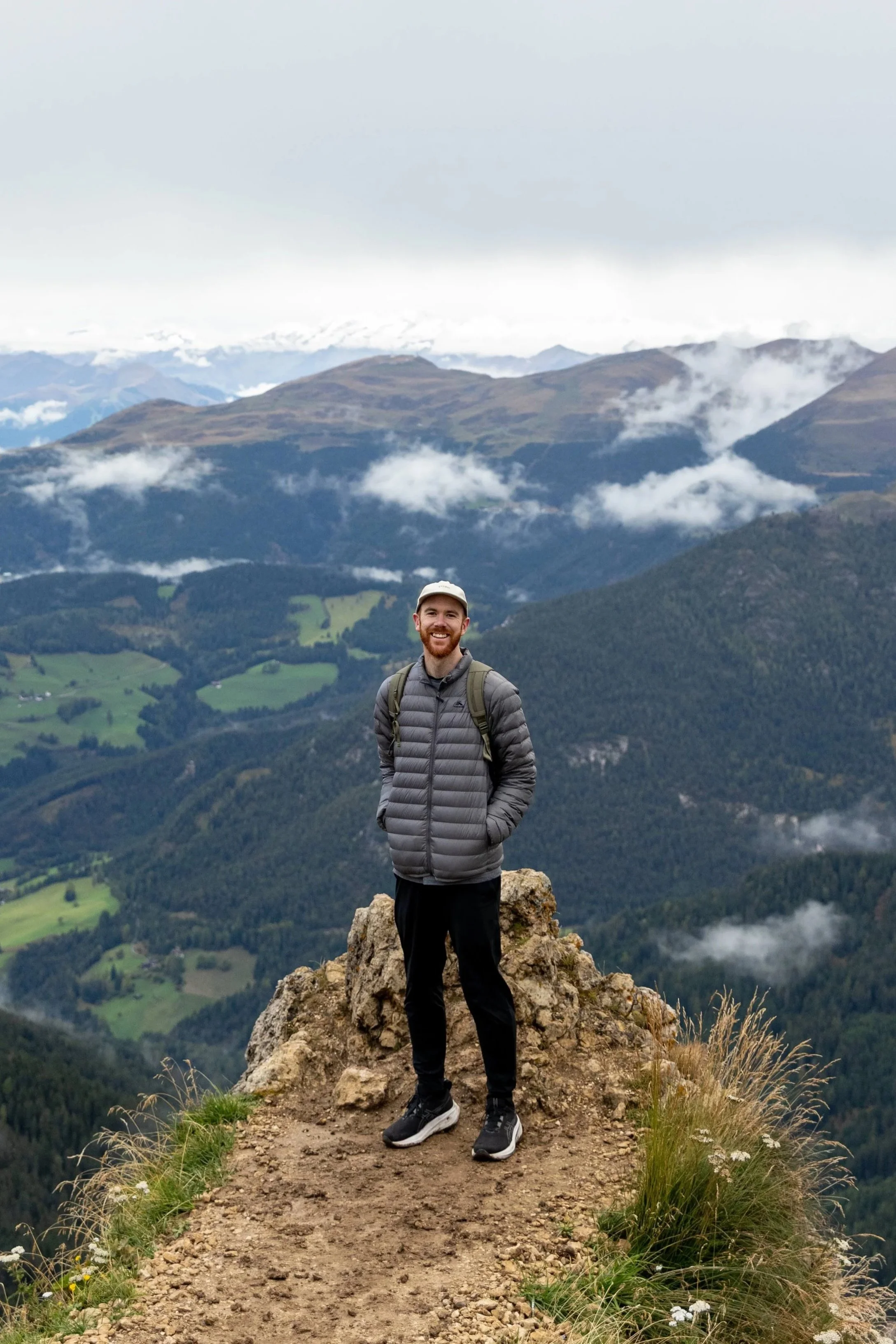 A man standing on a rocky trail at the top of a mountain ridge, wearing a gray jacket and black pants, with lush green mountains and clouds in the background.