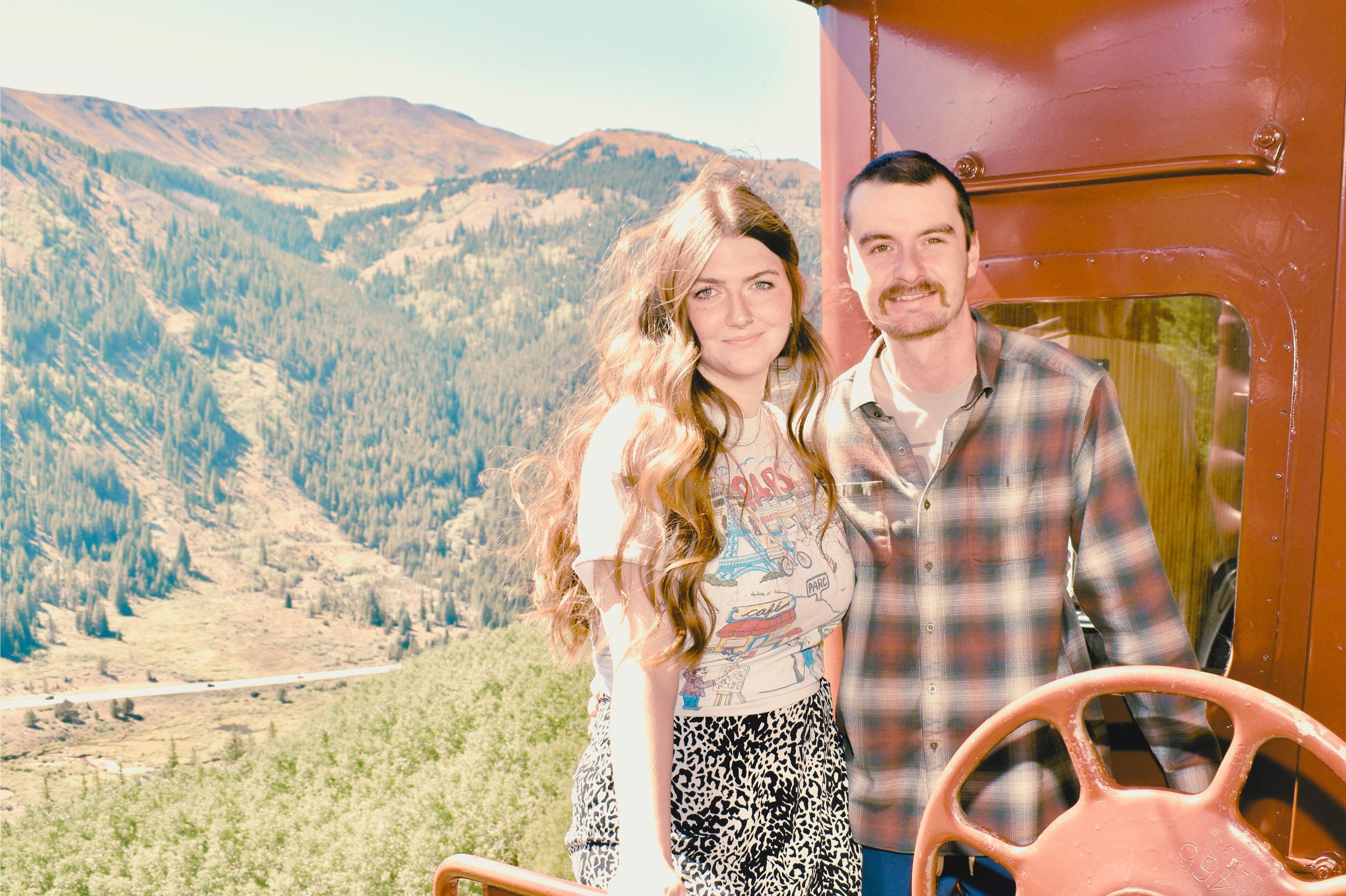 A young woman with long, wavy hair and a man with short dark hair and facial hair standing outside on a train, with lush green mountains and a valley in the background during daytime.