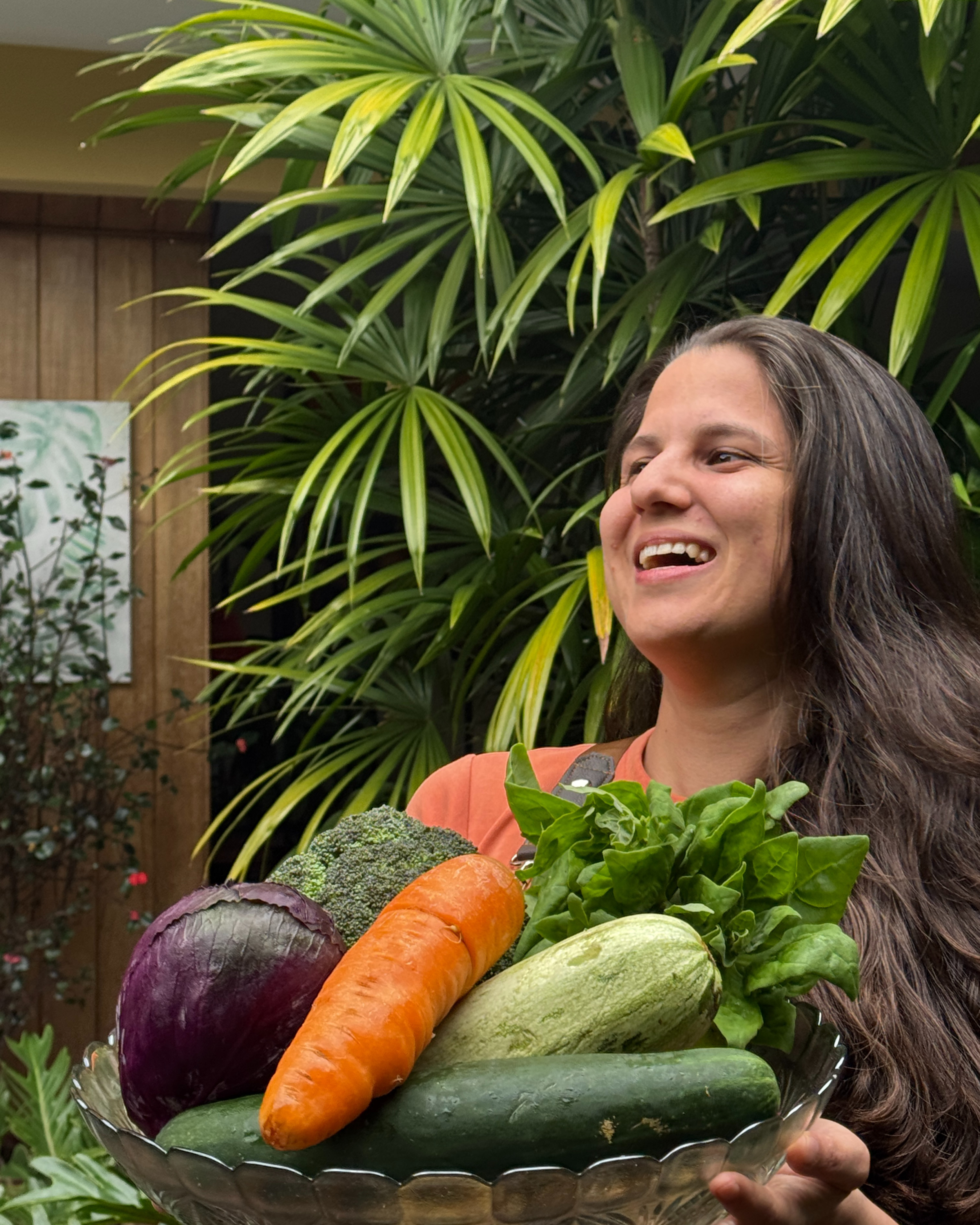 Mujer sonriendo sosteniendo un plato con varias verduras frescas, incluyendo coliflor, zanahoria, espinaca, pepino, calabacín y berenjena, en un entorno con plantas verdes de fondo.