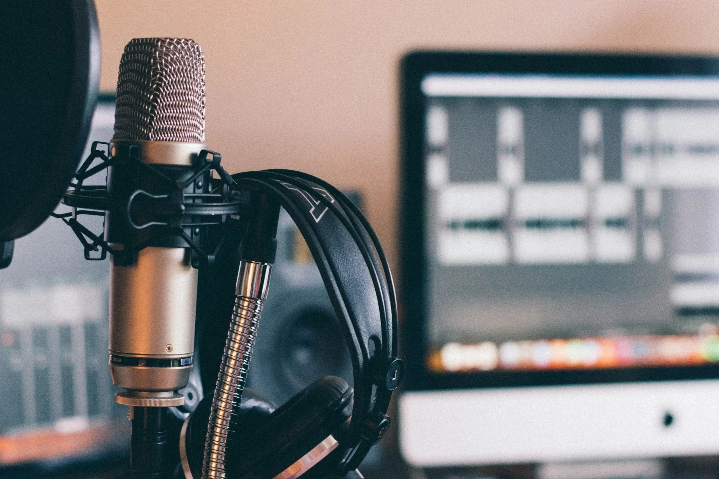 A professional microphone mounted on a stand, with headphones hanging on the same stand, positioned in front of a computer monitor displaying audio editing software.